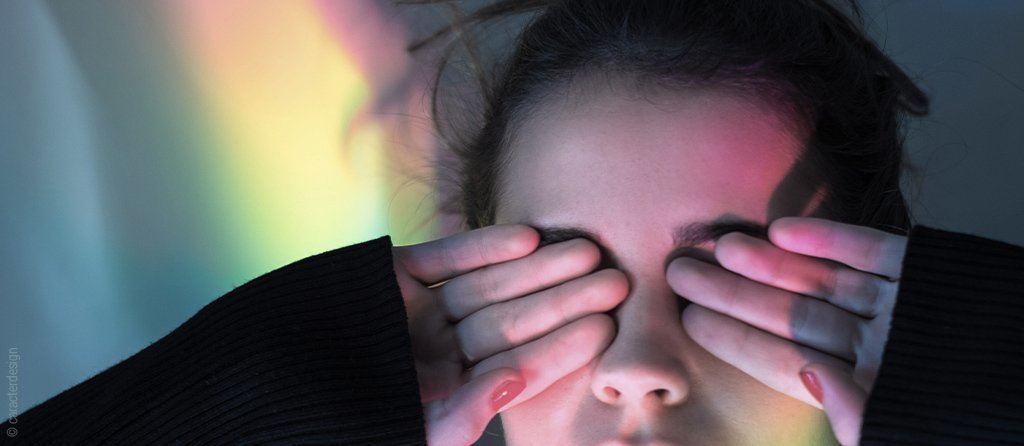 Foto: Eine Person hält sich die Finger vor die Augen. Neben ihr diffuses buntes Licht