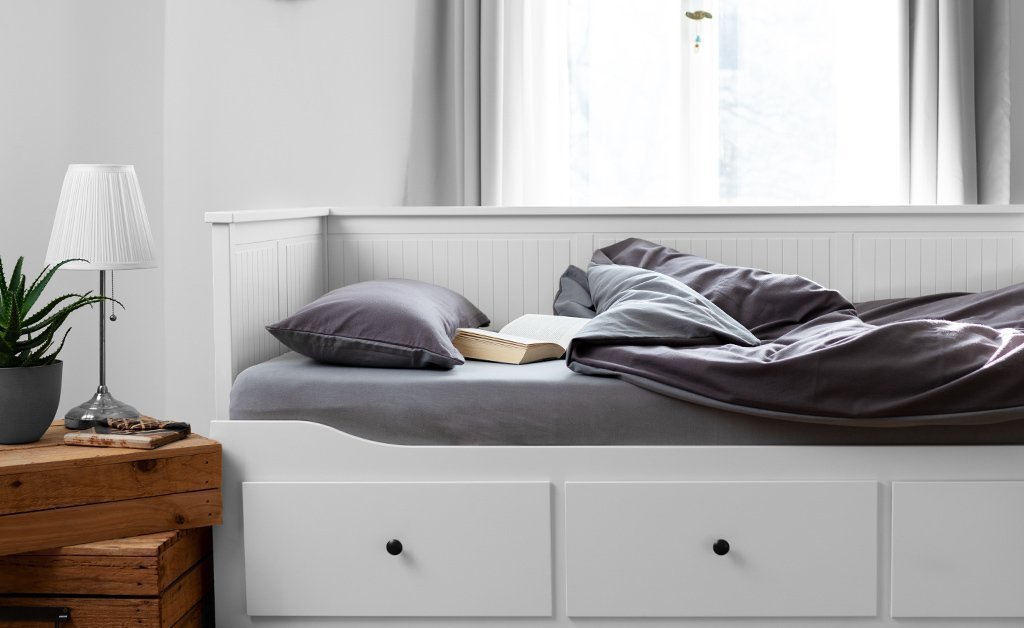 Foto: Links steht ein Beistelltisch, auf dem eine Lampe und eine Pflanze steht. Rechts daneben steht ein Bett, auf dem ein Buch liegt. Die Bettwäsche ist in einem grau und beigefarbenen Ton.
