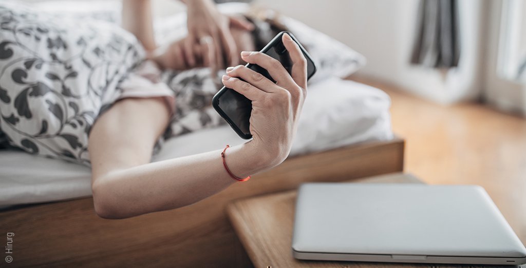 Foto: Eine Frau liegt im Bett und streckt ihren Arm in Richtung Nachttisch, auf dem ein Laptop liegt; sie hält ein Smartphone in der Hand – das alles widerspricht der Schlafhygiene.