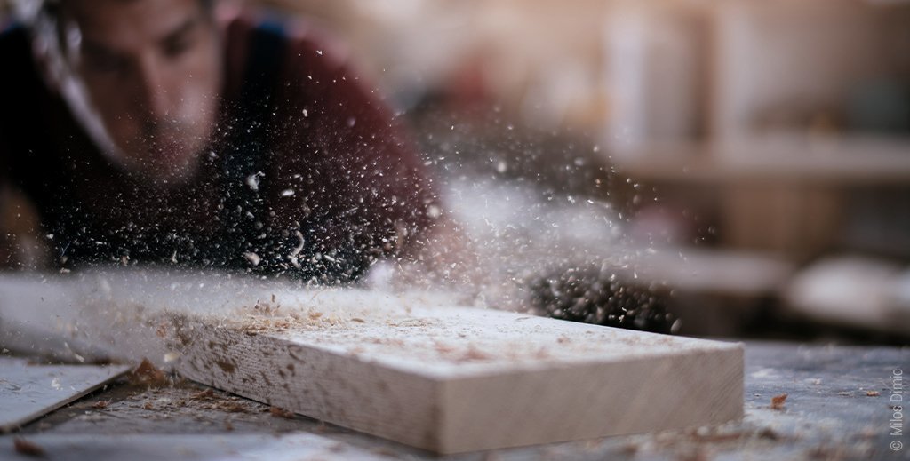 Foto: Ein Mann pustet Sägespäne von einer Holzlatte.