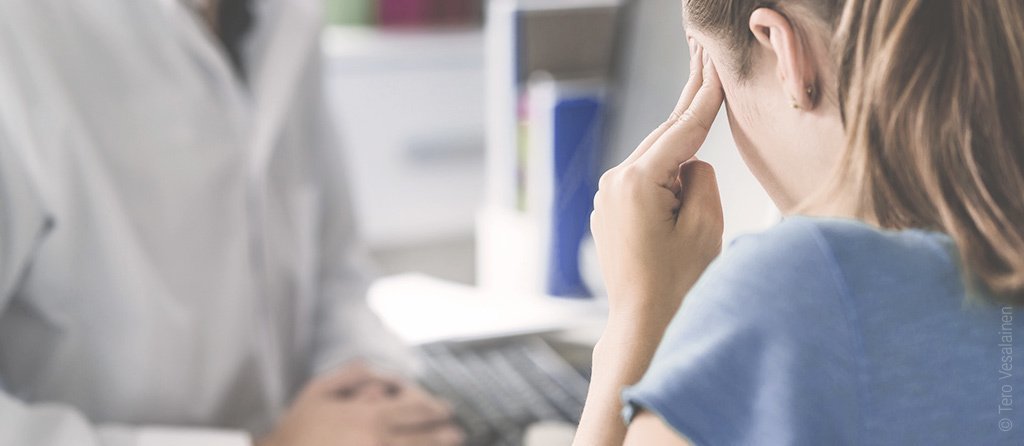 Foto: Im Vordergrund sitzt eine Person die sich die Schläfe reibt. Im Hintergrund sitzt eine Person im Kittel.