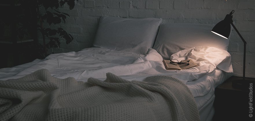 Foto: Ein Bett mit weißer Bettwäsche, daneben ein Nachttisch mit einer hell leuchtenden Nachttischlampe, die auf das ein Buch und Lesebrille auf dem Bett scheint.
