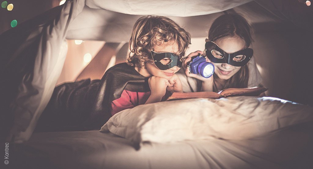 Foto: Zwei kostümierte Kinder lesen mit einer Taschenlampe ein Buch in ihrer selbst gebauten Höhle unter der Bett-Decke.