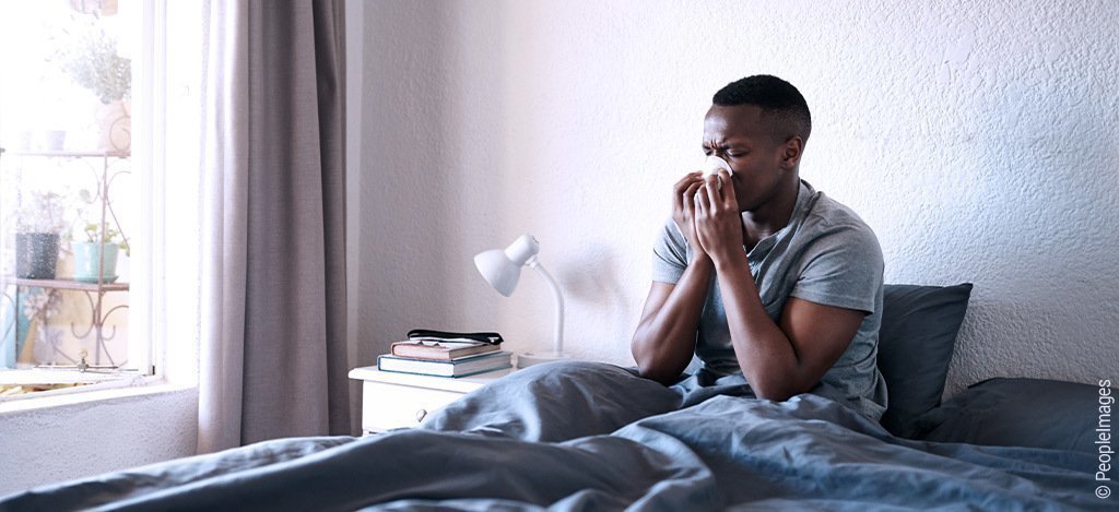 Foto: Ein Mann sitzt im Bett und schnäuzt in ein Taschentuch.