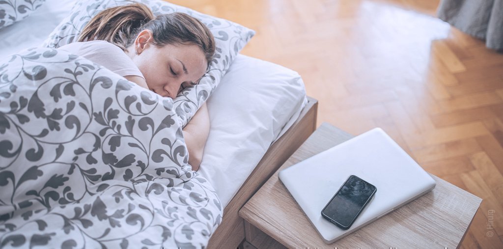 Foto: Eine Frau schläft im Bett. Auf dem Nachttisch neben ihr liegt ein Smartphone.