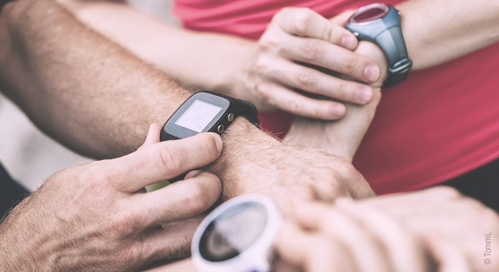 Foto: Drei Personen stehen im Kreis. Der Bildausschnitt fasst nur die Unterarme mit Fitnessarmbändern am Handgelenk und die jeweils andere Hand, die einen Knopf am Armband drückt.