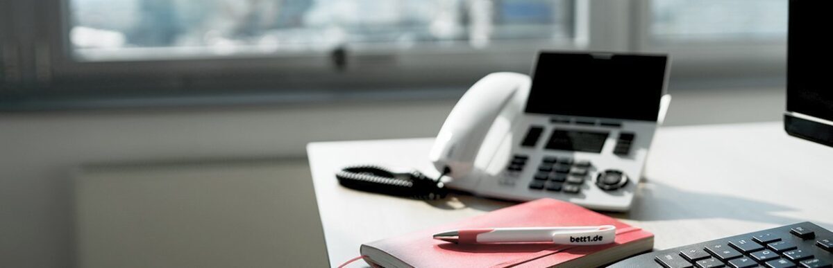 Foto: Arbeitsplatz im Büro samt Telefon, Computer und Notizbuch mit Stift