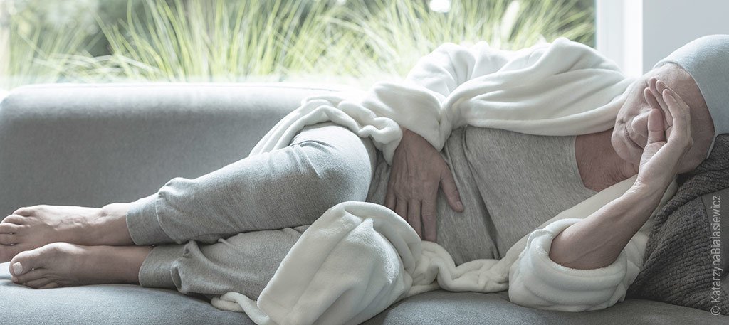 Foto: Krebspatientin liegt gekrümmt auf dem Sofa, hält sich eine Hand vor den kahlen Kopf und mit der anderen auf den Bauch.