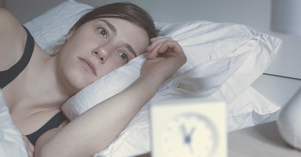 Foto: Eine Person liegt im Bett und starrt ins Leere. Der Wecker im Vordergrund zeigt 1 Uhr nachts.