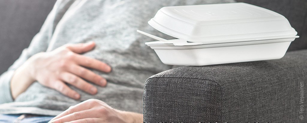 Foto: Eine Person sitzt auf dem Sofa und hält sich den Bauch. Im Vordergrund liegt eine leere halbgeöffnete Essen-To-Go-Verpackung.