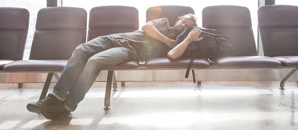 Foto: Ein Mann schläft am Flughafen, sein Kopf liegt auf seiner Reisetasche.