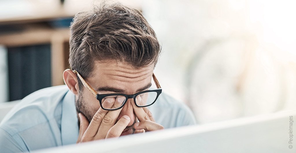 Eine Person sitzt vor einem Monitor. Der Kopf ist auf die Hände gestützt, die Finger unter der Brille reibend auf den Augenlidern