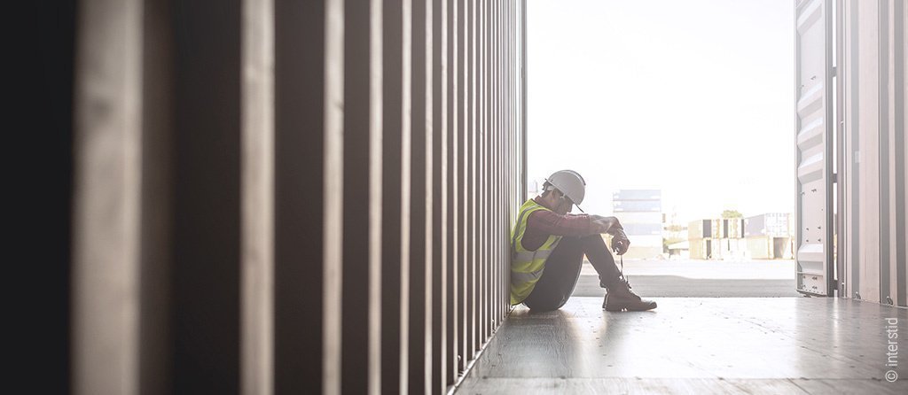 Foto: Ein Bauarbeiter sitzt auf einem Container-Boden und hält den Kopf gesenkt.