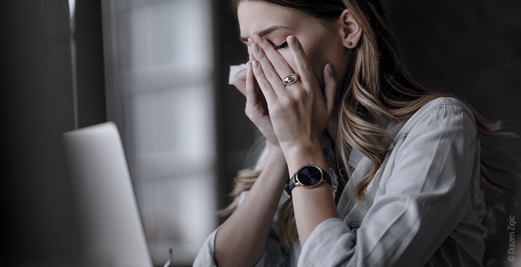 Foto: Eine Persoon sitzt aufrecht am Schreibtisch und vergräbt ihr Gesicht in den Händen