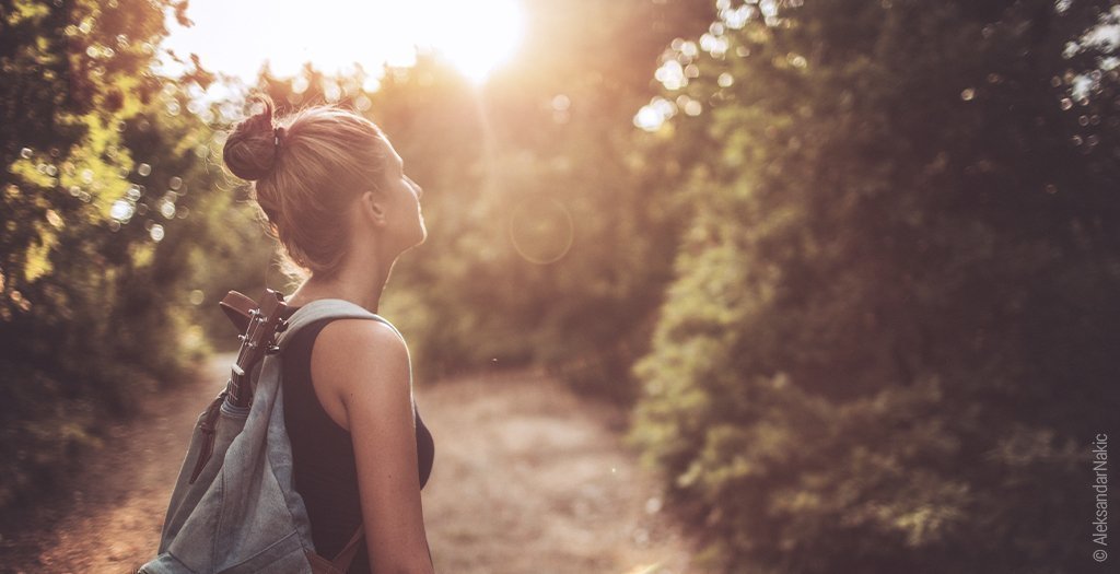 Foto: Eine junge Person mit Rucksack steht auf einem Waldweg und schaut in die orange Sonne.