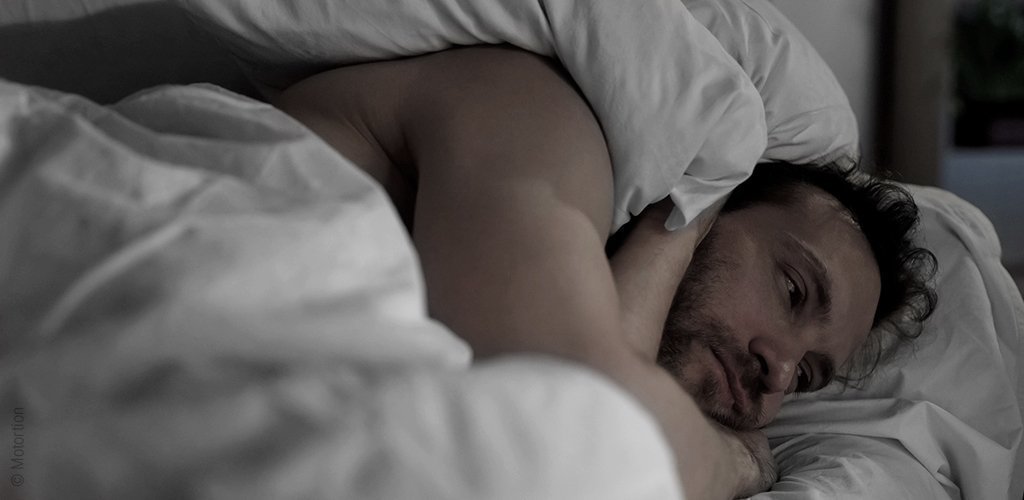 Foto: Ein sichtlich gestresster Mann liegt im Bett; das Hormon Cortisol kann uns vom Schlafen abhalten. 