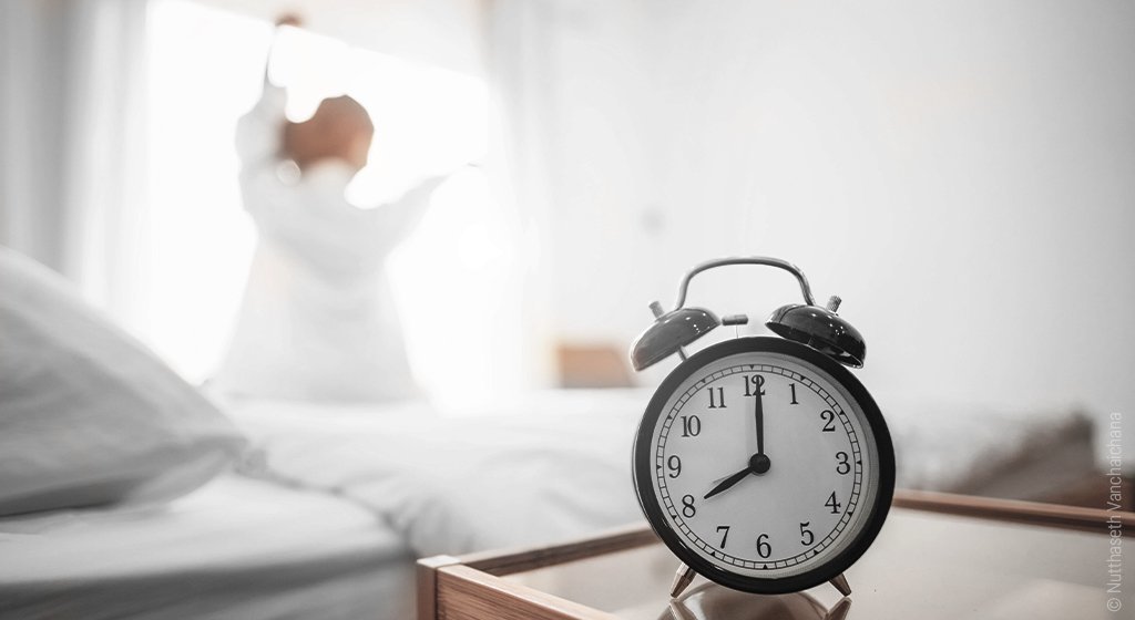 Foto: Im Vordergrund ein Wecker, im Hintergrund sitzt eine Frau auf dem Bett und streckt sich; es geht um das Schlafhormon Serotonin.