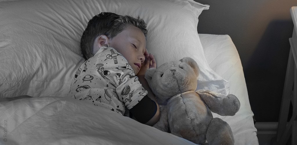 Foto: Ein Kind liegt neben einem Teddybären im Bett und schläft; das Schlafhormon Somatropin fördert das Zellwachstum.