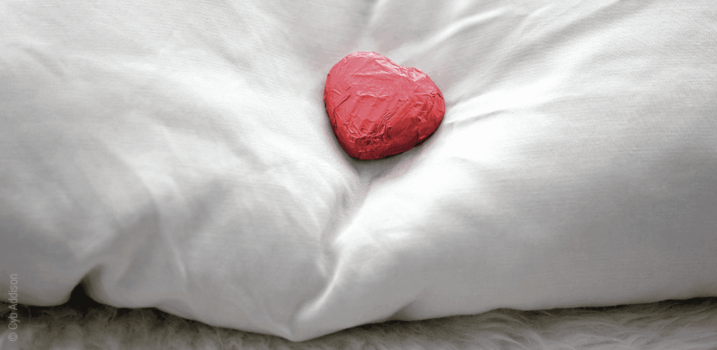 Foto: Das essbare Betthupferl – ein in roter Alufolie eingewickeltes Schokoladenherz liegt auf einem Kopfkissen.