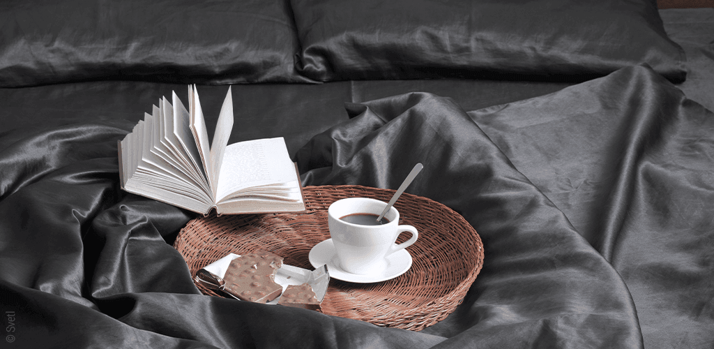 Foto: Ein Strohtablett mit Espresso und einer Tafel Schokolade steht auf einem Bett, daneben ein aufgeschlagenes Buch.