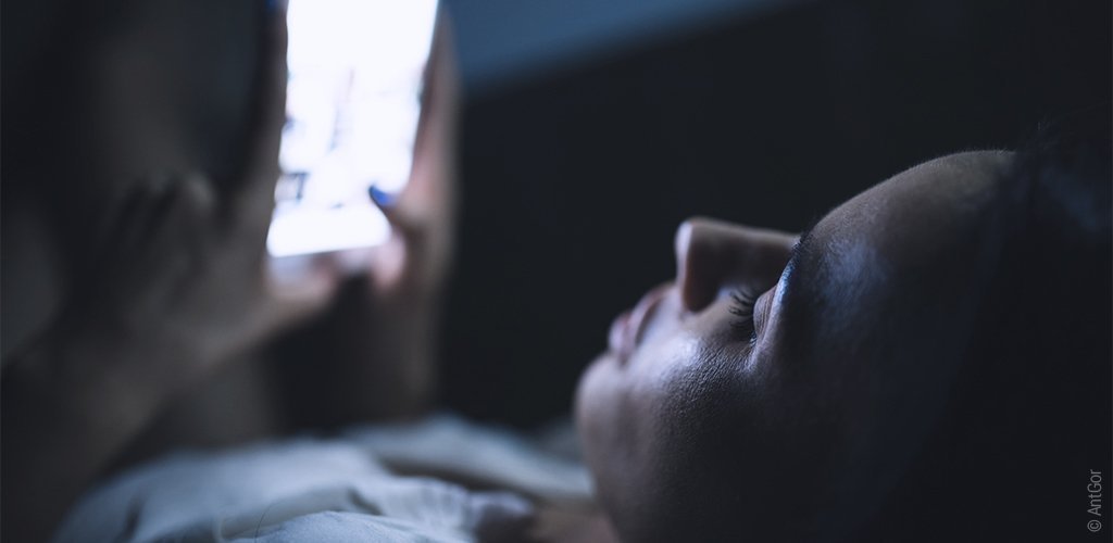 Foto: Eine Frau liegt im Bett und guckt dabei auf ihr Handy.