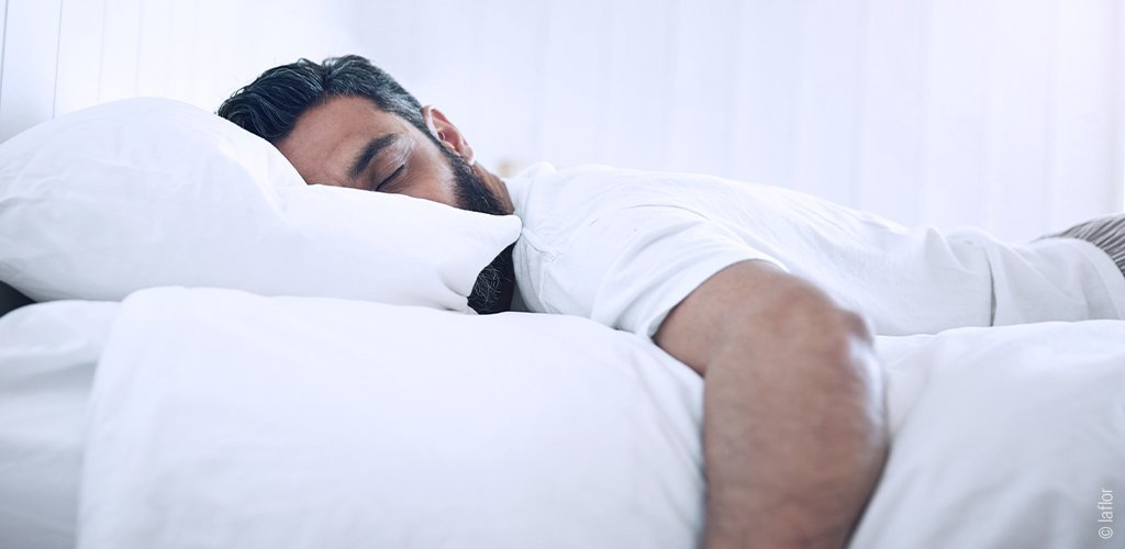 Foto: Ein Mann liegt im Bett und schläft, sein Arm hängt an der Bettkante hinunter.