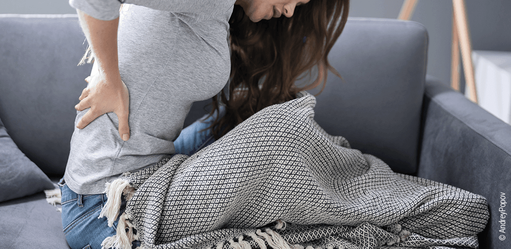 Foto: Eine Frau sitzt aufrecht auf einem Sofa und hat ihre Hand am Rücken.