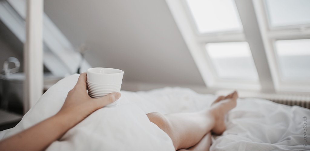 Foto: Eine Frau liegt im Bett und sieht aus dem Fenster. Sie hat eine Tasse in der Hand.