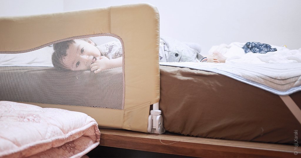 Foto: In einem großen Bett liegt ein lachendes Kleinkind. Es schaut durch ein klappbares Bettgitter, das mit Netz bespannt und unter der Matratze festgesteckt wurde, aus dem Bett