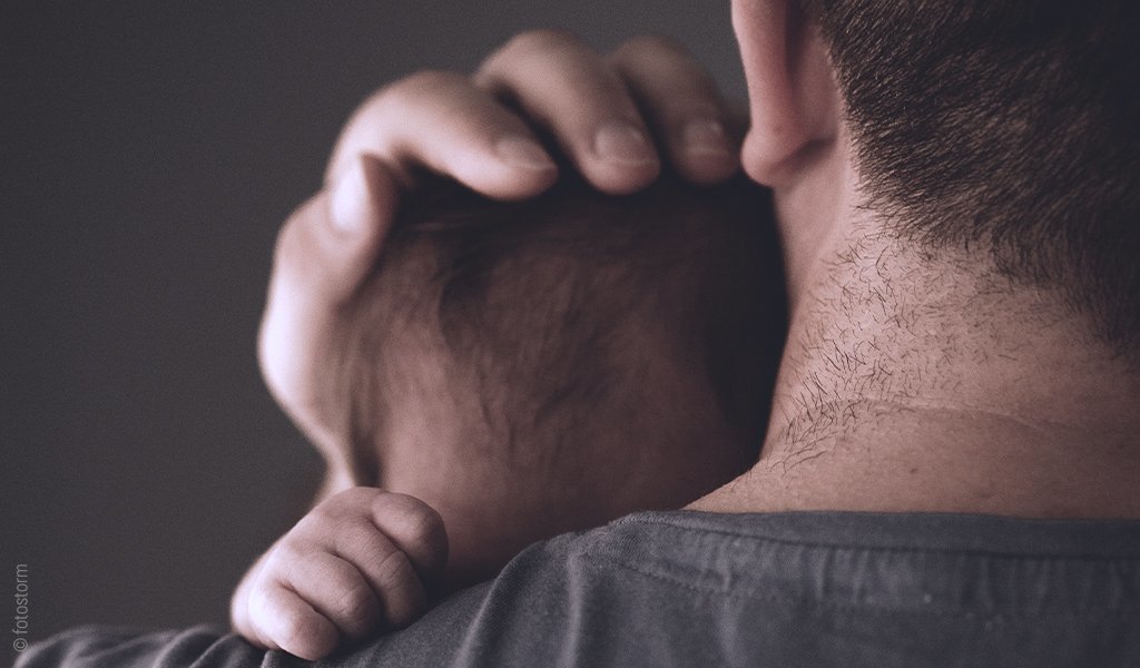 Foto: Eine Person von hinten. Ein Babykopf ist gegen die Schulter der Person gestützt, die Hand des Erwachsenen hält den Hinterkopf. Die Babyhand greift in den Stoff des Shirts an der Schulter.