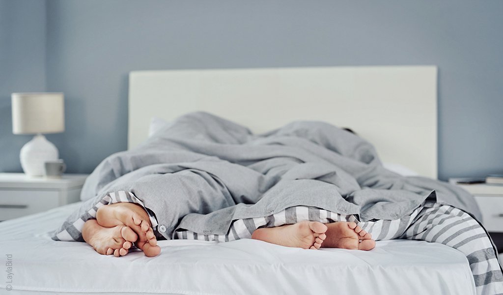 Foto: Zwei Personen liegen in einem Bett. Nur ihre Füße schauen unter der Decke hervor.