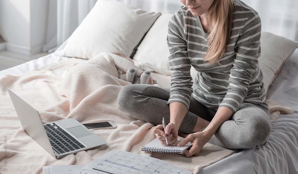 Foto: Eine Person sitzt im Bett und schreibt Notizen, vor ihr ein Laptop.