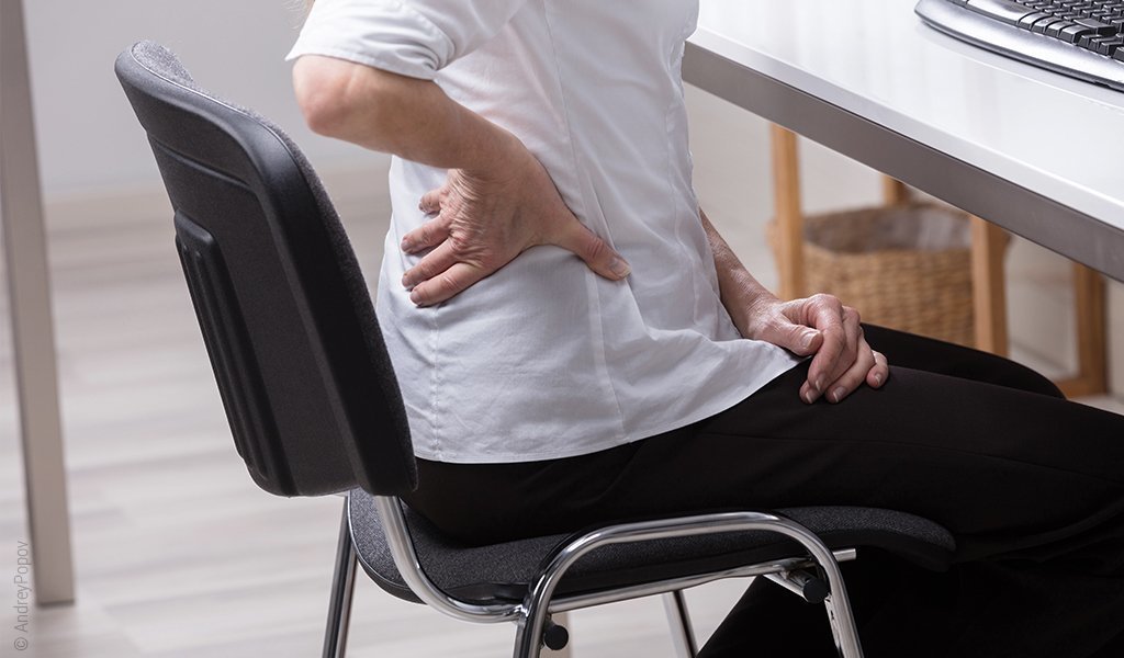 Foto: Eine Person sitzt auf einem Bürostuhl und fasst sich an den unteren Rücken im Bereich der Lordose.