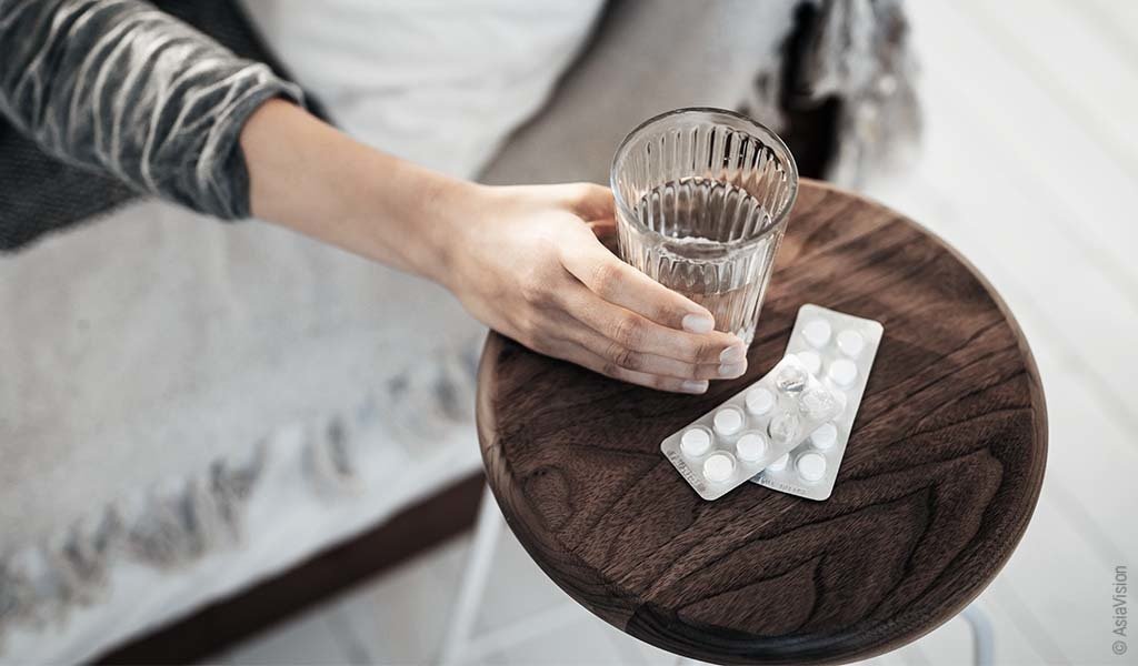 Foto: Eine Hand greift zu einem Glas Wasser; daneben liegen zwei Blister mit Tabletten.