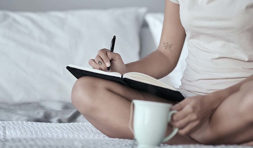 Foto: Eine Person sitzt mit einer Kaffeetasse in der Hand auf dem Bett und schreibt in ein Buch.