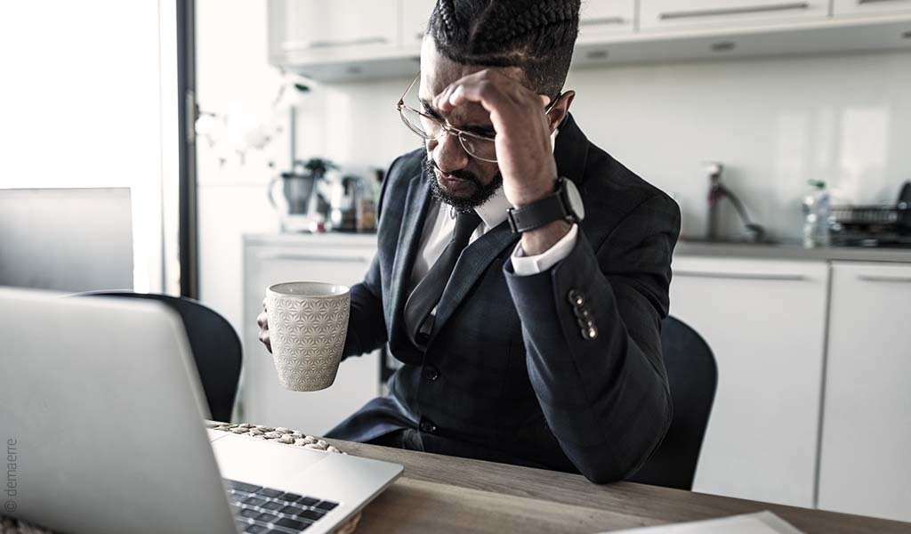 Symbolfoto Insomnie: Eine Person sitzt mit Kaffeetasse vor einem Laptop und hält sich den Kopf.