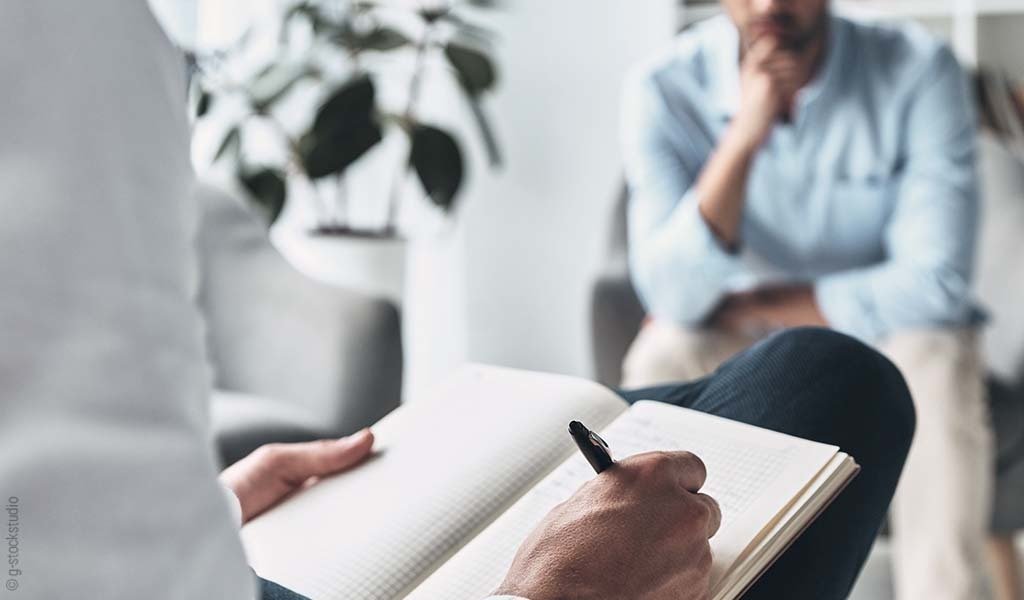 Foto: Zwei Personen sitzen sich gegenüber; die eine schreibt in ein Buch.