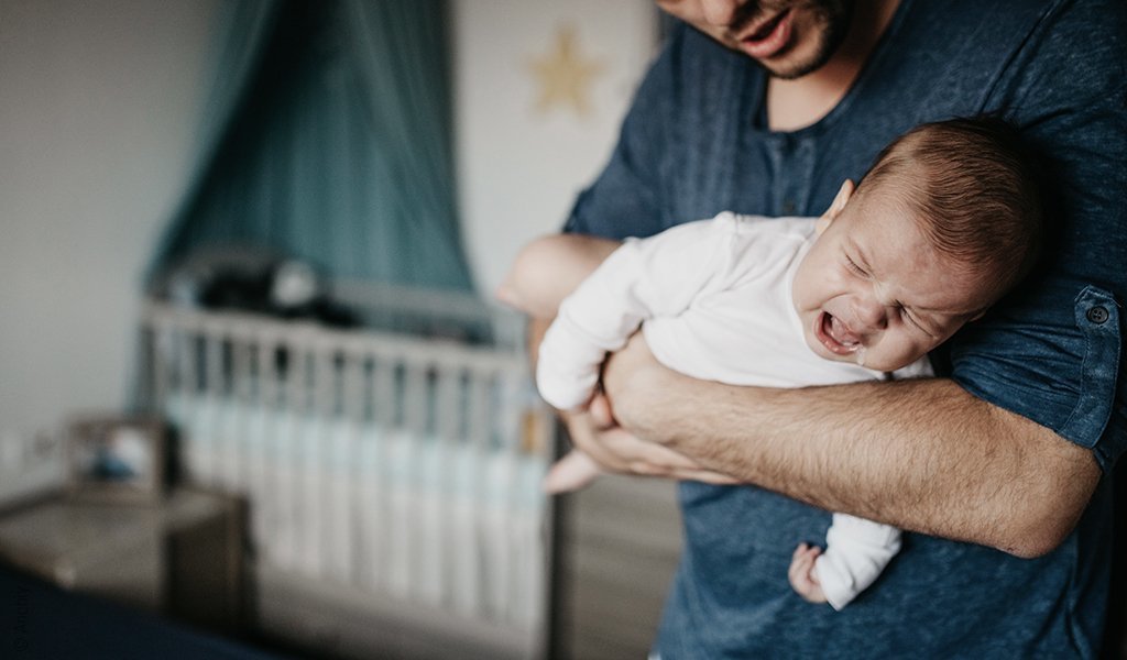 Foto: Eine Person hält ein schreiendes Baby im Fliegergriff. Dahinter unscharf das Kinderzimmer mit Gitterbett.