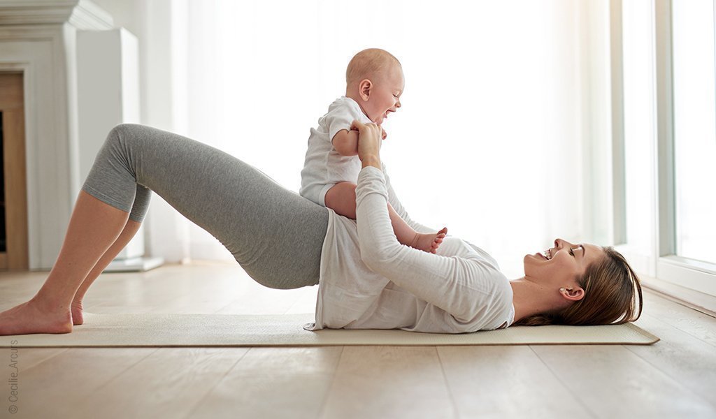 Foto: Ein Baby sitzt auf einer Person, die auf einer Sportmatte liegt und eine halbe Brücke macht. Beide schauen sich lachend an.