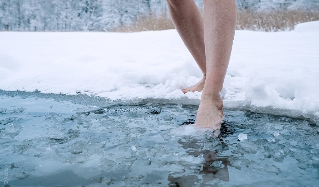 Foto: Eine Person streckt bei Schnee und Eis einen Fuß in einen eiskalten See.