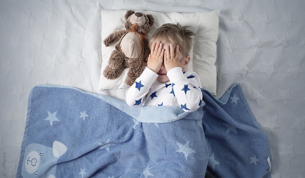 Foto: Ein Kind in Rückenlage unter einer Wolldecke und hält sich die Hände vors Gesicht. Neben ihm auf dem Kopfkissen liegt ein Teddybär.