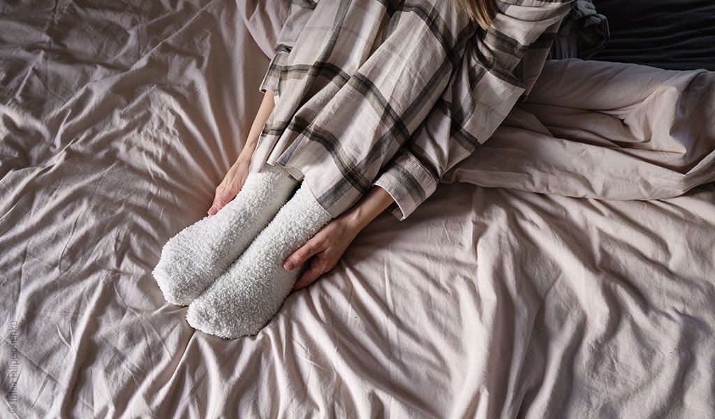 Foto: Eine Person sitzt im Bett und hat Kuschelsocken an; sie umklemmt ihre Hände an ihren Füßen.