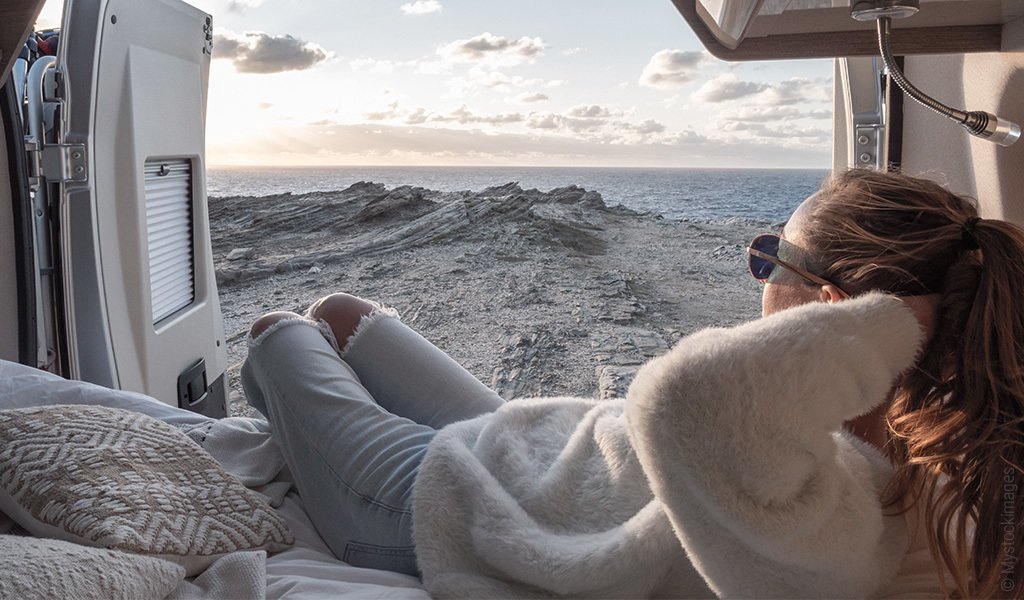 Foto: Eine Person sitzt im Wohnwagen und schaut sich bei geöffneter Tür die Landschaft an.