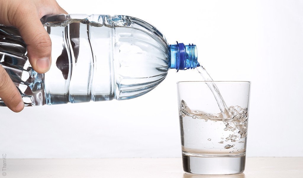 Foto: Nahaufnahme einer Hand, die Wasser aus einer Plastikflasche in ein Glas gießt.