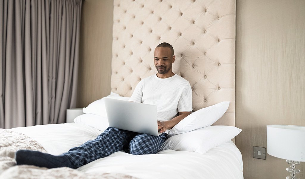 Foto: Eine Person sitzt mit Laptop auf einem Bett und lehnt sich an die Bett-Rückwand an.