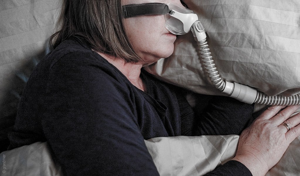 Foto: Eine Person liegt im Bett und trägt eine CPAP-Beatmungsmaske auf der Nase.