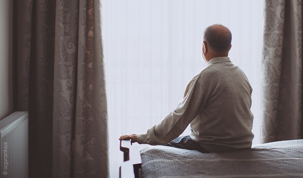 Foto: Eine Person sitzt, den Betrachtenden den Rücken zugewandt in gerader Haltung auf einem Bett und schaut aus dem Fenster.