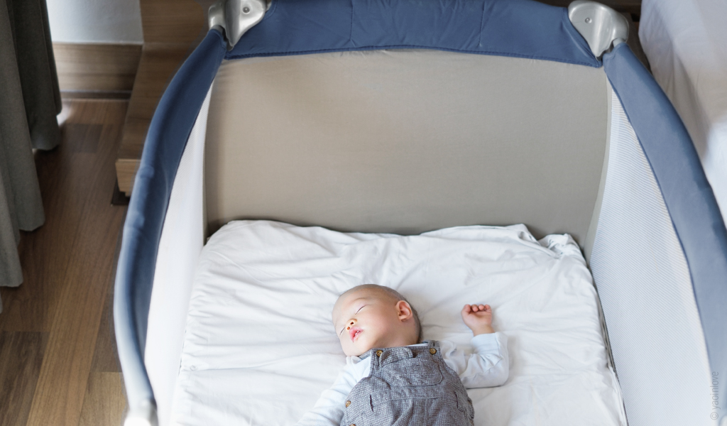 Foto: Blick in ein Reisebett mit Babymatratze 60x120 cm in dem ein Baby auf seinem Rücken schläft.