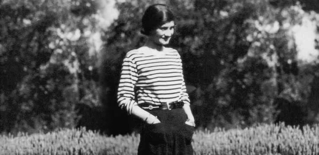 Foto: Ein schwarz-weiß Foto der Modedesignerin Coco Chanel in jüngeren Jahren; sie trägt ein quer gestreiftes Oberteil und eine legere schwarze Hose.