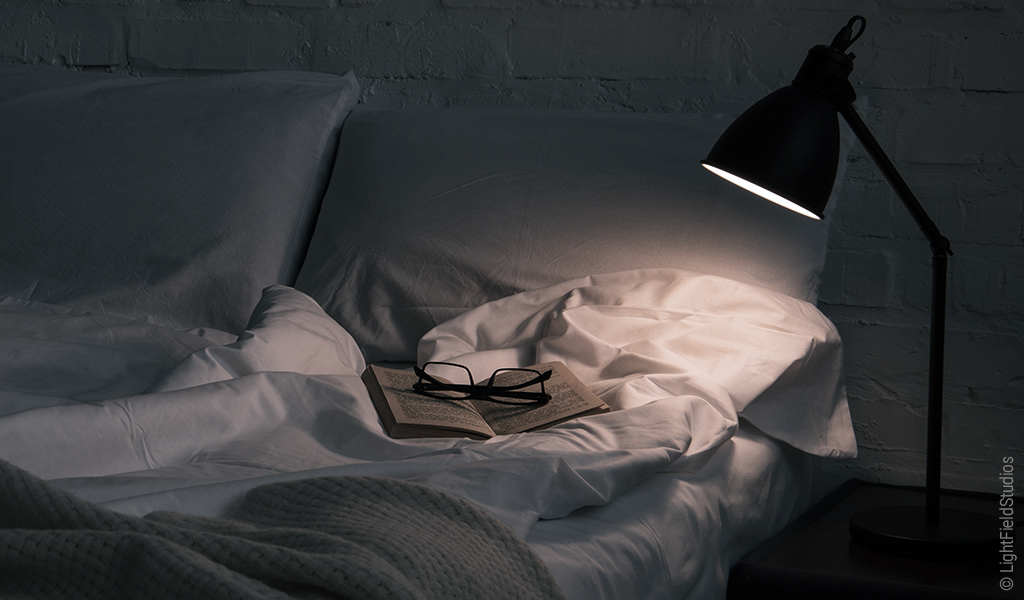 Foto: Eine Stehlampe beleuchtet einen kleinen Teil eines Betts. Darauf liegt ein aufgeschlagenes Buch und eine Brille.